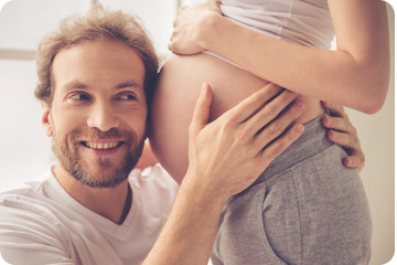 Un homme colle ton oreille contre le ventre de sa femme enceinte et entoure son ventre de ses mains, il sourit.